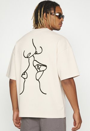 Mann trägt beige oversized T-Shirt mit schwarzer abstrakter Linienzeichnung von zwei Gesichtern auf dem Rücken, graue Hose und dunkle Sonnenbrille.
