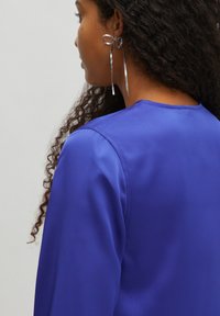 Koninklijkblauwe satijnen top met een gladde textuur. Lange, licht uitlopende mouwen. Metallic oorbellen met een strikontwerp en lange, slanke druppels.