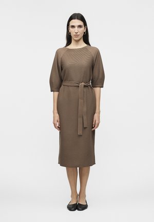 Γυναίκα με μακριά σκούρα μαλλιά που φοράει ένα καφέ φόρεμα μέχρι το γόνατο με μανίκια τρία τέταρτα και ζώνη δεμένη στη μέση, στέκεται μπροστά σε λευκό απλό φόντο.