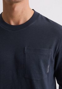 Granatowa koszula z długim rękawem wykonana z teksturowanej tkaniny, z okrągłym dekoltem i kieszenią na piersi z małą metką z boku.