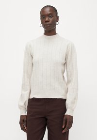 Maglione lavorato a maglia di un beige chiaro con colletto alto, texture a costine verticali e maniche lunghe a sbuffo, abbinato a pantaloni marrone scuro.