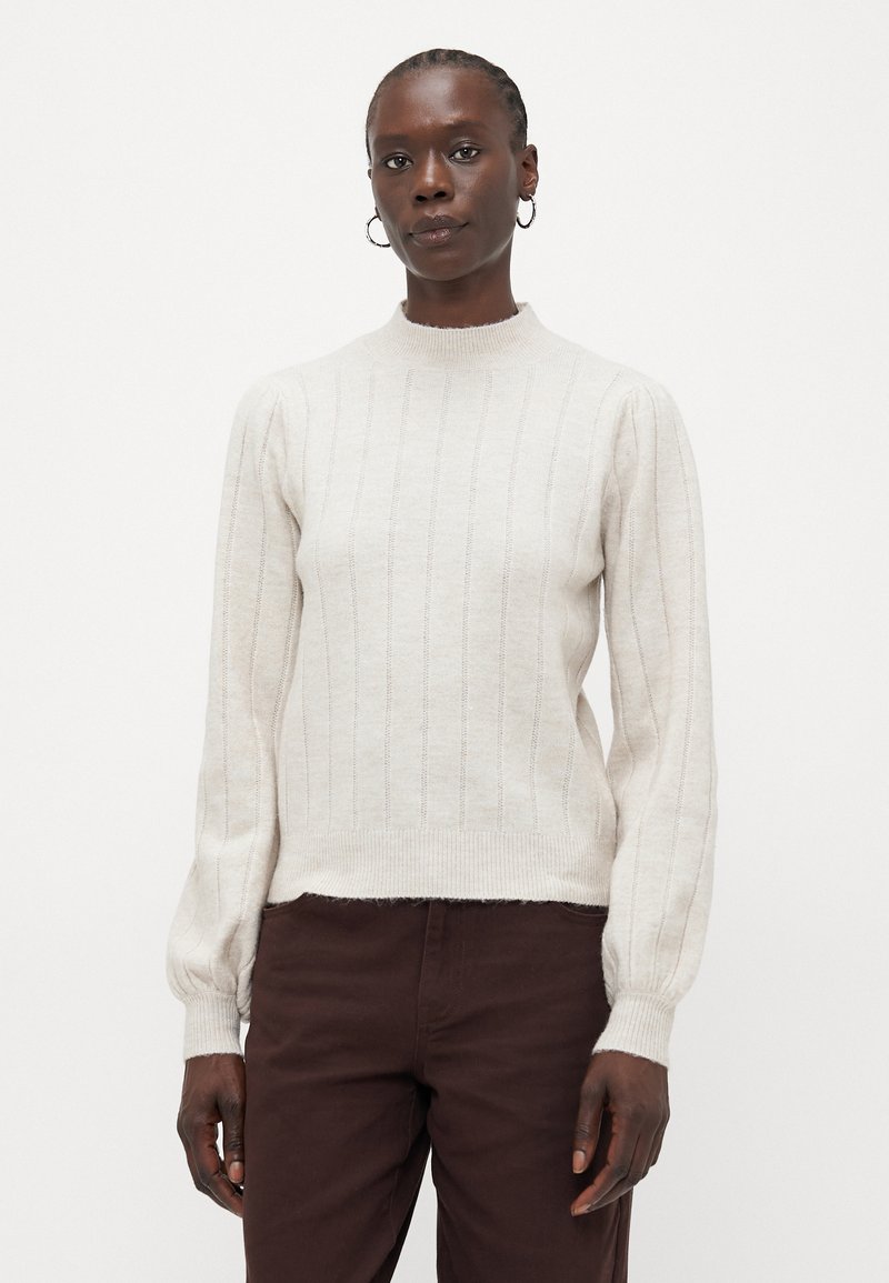 Maglione lavorato a maglia di un beige chiaro con colletto alto, texture a costine verticali e maniche lunghe a sbuffo, abbinato a pantaloni marrone scuro.