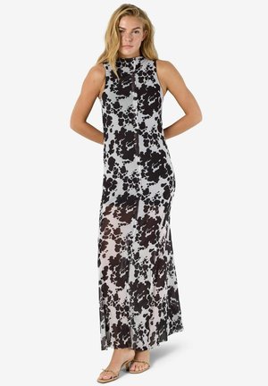 Kvinne iført ermeløs, hellang kjole med svarte og grå abstrakte flekker, stående med hendene bak ryggen, lette hæler.