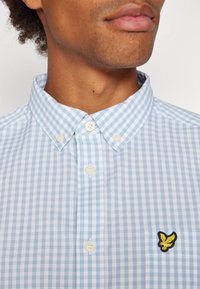 Light blue and white checked shirt with a button-down collar, featuring white buttons and a yellow embroidered logo on the chest pocket.