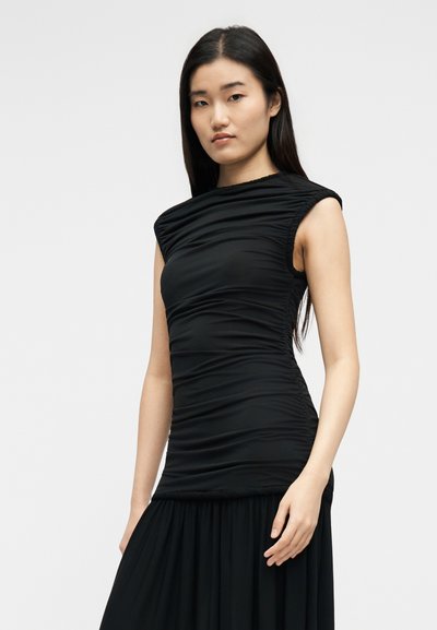 Νεαρή γυναίκα με μακριά σκούρα μαλλιά, ντυμένη με αμάνικο μαύρο φόρεμα με ρίγες, μπροστά σε απλό φωτεινό φόντο.