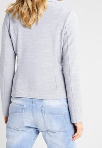 Blazer gris clair à côtelés avec une silhouette ajustée, des manches longues et un col haut, associé à un jean en denim bleu clair avec des poches arrière.