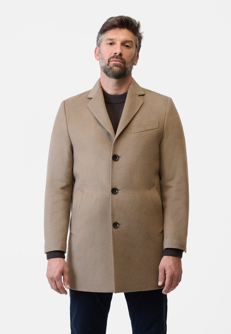 Beige ullkappa med spetsig krage, enkelknäppt design och tre framknappar. Har två sidofickor och en bröstficka.