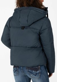 Dunkelblaue gesteppte Jacke mit abnehmbarem Kapuzen, gestepptem Design und einem kleinen Logo-Tag auf der Rückseite; kombiniert mit blauen Jeans mit detaillierten Taschen.