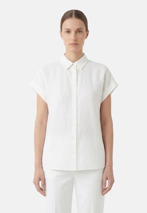 Donna con capelli tirati all'indietro che indossa una camicia bianca a maniche corte con bottoni e pantaloni bianchi, in piedi davanti a uno sfondo semplice.