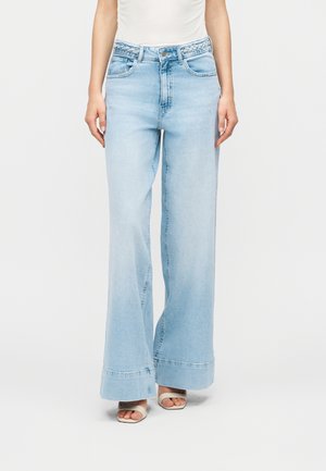 Nő világoskék, széles szárú farmert visel, amelynek derékrészén fonott szalag található, fehér, nyitott orrú szandállal párosítva, egyszerű háttér előtt áll.