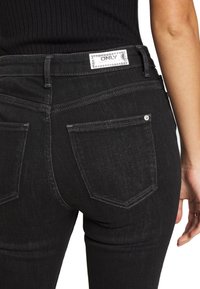 Los pantalones vaqueros de mezclilla negra presentan una textura suave, un diseño de cinco bolsillos, con una etiqueta de marca en la parte posterior de la cintura y costuras en contraste.