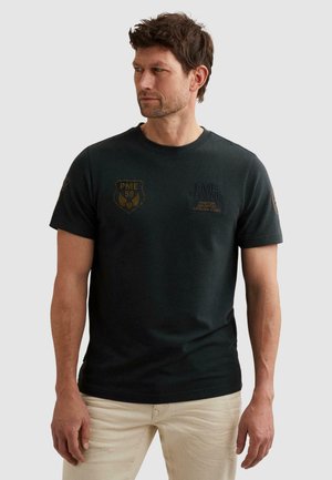 Man wearing black PME Legend short-sleeve shirt with logos, beige pants, looking to the side against plain light background.