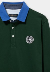 Dunkelgrünes Polohemd mit blauem Kragen, einer weißen Logoplakette auf der Brust und drei Knöpfen am Hals. Glatte Stoffstruktur.