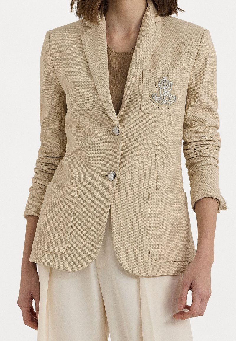 Blazer beige ajusté avec deux poches avant, écusson brodé sur la poche poitrine gauche, porté sur un haut beige assorti et un pantalon crème.
