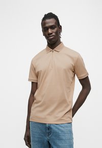 Νεαρός με σκουρόχρωμο δέρμα και κοτσίδες, που φοράει μπεζ κοντομάνικο πόλο με φερμουάρ και μπλε τζιν, μπροστά σε λευκό φόντο.