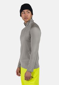 Man som bär svart mössa, grå långärmad zip-up-tröja och neongula byxor, står sidledes och tittar åt vänster.