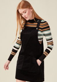 Musta corduroy-sali, jossa on kaksi etutaskua ja säädettävät hihnat, käytetty monivärisen raidallisen pitkähihainen paidan päällä.