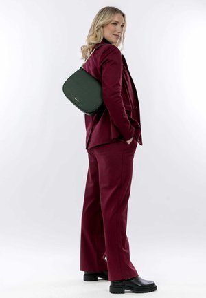 Grüne Leder-Handtasche mit runder Form und strukturiertem Oberflächen, über der Schulter getragen. Modell in einem burgunderfarbenen Anzug mit Schlaghose.