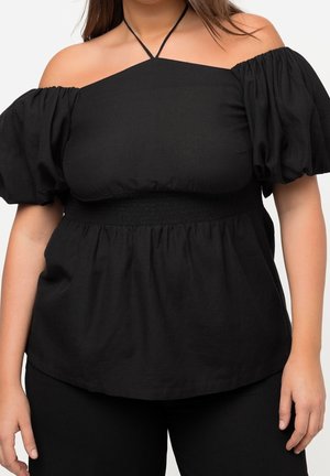 Kvinde iført en sort off-shoulder top med puffede ærmer og halterneck, kombineret med sorte bukser.
