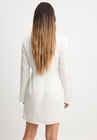 Žena s dlouhými ombré vlasy, oblečená v bílé dlouhé šaty po kolena s dlouhým rukávem, stojící čelem k jednobarevnému bílému pozadí.