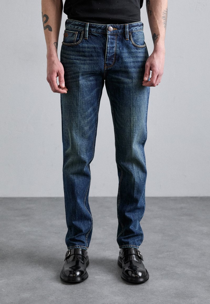 Tamsiai mėlynos džinsinės kelnės su klasikinio kirpimo, su švelniu išblukimu, tradiciniu penkių kišenių dizainu ir kontrastingomis siūlėmis.