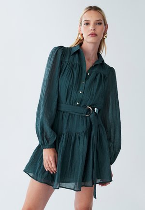 Diep groene jurk van textuurstof, met een knopenvoorzijde, lange pofmouwen, een ceintuur in de taille en een uitlopende rok.