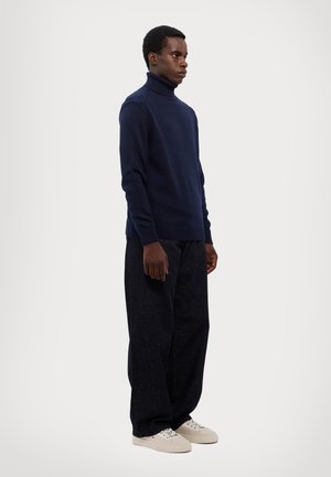 Maglione a collo alto di colore blu navy in materiale morbido, abbinato a pantaloni in denim scuri e larghi e sneaker di colore chiaro. Design semplice e moderno.