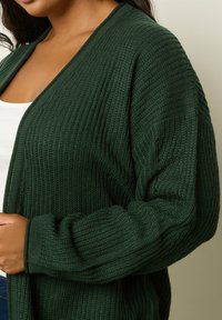 Kvinna som bär en mörkgrön chunky stickad cardigan över en vit skjorta, visad från bröstet till midjan mot en beige bakgrund.