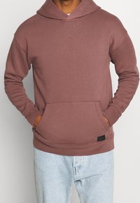 Bordowa bluza z kapturem z kieszenią kangura, długimi rękawami i sznurkiem przy kapturze. Wykonana z miękkiej tkaniny z ściągaczami na mankietach oraz małą metką z logo.