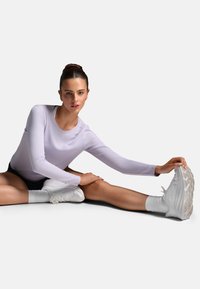 Žena v lila tričku s dlhým rukávom a čiernych šortkách sedí a naťahuje sa, predkláňa sa, aby sa dotkla svojej bielej tenisky na vystretej nohe.