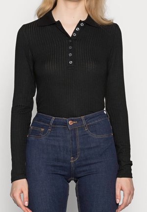 Kvinde iført en tætsiddende sort langærmet ribbet polo med knapper og højtaljede mørkeblå jeans mod en hvid baggrund.