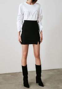 Žena ve bílé dlouhé rukávové topu s řasením, černé úzké minisukni s předním rozparkem a černých kozačkách na podpatku do poloviny lýtek stojí na betonové podlaze.