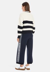 Strickpullover in Creme- und Marineblau-Streifen, mit lockerer Passform. Kombiniert mit marineblauen Hosen mit weißen Seitenstreifen und grafischem Text, ergänzt durch weiße Sneaker.