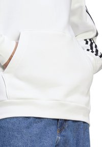 Sudadera blanca con un gran bolsillo delantero, con acentos de rayas negras a lo largo de las mangas. Tejido suave con una textura lisa.