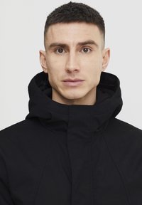 Sort hættejakke med en struktureret design, der har en høj krave, synlige sømme og en glat, mat overflade.