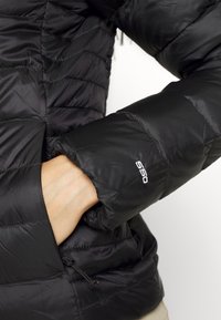 Main dans la poche d'une veste noire matelassée avec "550" brodé sur la manche, montrant une partie de l'avant et des détails de la manche de la veste.