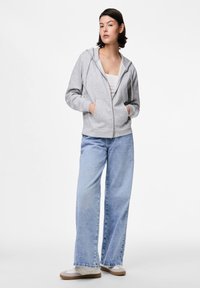 Sivi zip-up dukserica s mekanom teksturom, prednjim džepovima i kapuljačom, u kombinaciji sa svijetloplavim širokim trapericama i bijelim tenisicama.