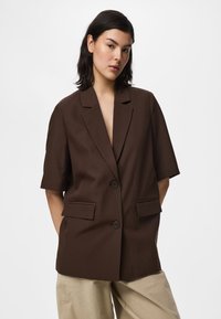 Νέα γυναίκα με σκούρα μαλλιά φοράει καφέ σακάκι με κοντά μανίκια πάνω από μπεζ παντελόνια, στέκεται με τα χέρια στις τσέπες μπροστά σε ένα απλό φόντο.