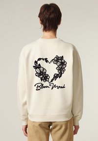 Felpa color crema con un design di cuore floreale nero e testo "Bloom Mood" ricamato sulla parte posteriore. Vestibilità comoda e polsini a costine.