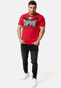 Sarkans kokvilnas T-krekls ar galvaskausa grafiku un tekstu "TAPOUT SIMPLY BELIEVE." Apvienots ar melniem džinsiem un baltiem sporta apaviem.
