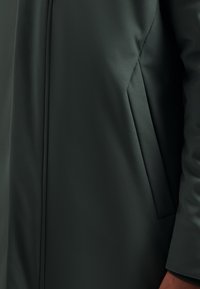 Donkergroene jas met een gladde textuur, verticale naden en een zijzak. Het ontwerp is afgestemd op een gestructureerde pasvorm.