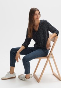 Marineblau gemustertes Langarm-Bluse mit gerafften Details, kombiniert mit dunkelblauen Jeans und weißen Sneakern, sitzt auf einem Holzstuhl.