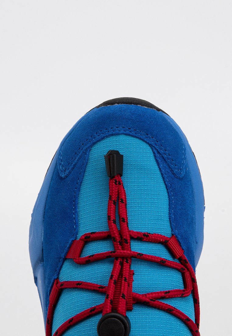 Chaussure de sport bleue avec une tige en daim et en textile, dotée d'un système de laçage rouge et d'une semelle extérieure noire. Texture lisse et forme arrondie.
