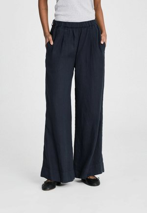 Személy bő, széles szárú fekete nadrágot visel gumis derékrésszel, fekete balettcipővel és világosszürke felsővel párosítva.