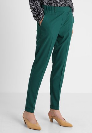 Kobieta ubrana w zielone spodnie zwężane z mankietami, zestawione z beżowymi mule na niskim obcasie z zaostrzonym noskiem oraz bluzką w czarno-biały wzór.