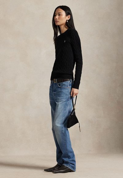 Mujer de pie con suéter de punto tipo cable negro, jeans azules, zapatos negros puntiagudos, sosteniendo un pequeño bolso negro sobre fondo neutro.
