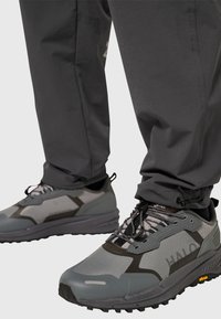 Grijze sportschoenen met mesh- en rubberaccenten, voorzien van een robuuste zool en zwarte veters, gecombineerd met grijze broek die ter hoogte van de enkels is vastgemaakt.