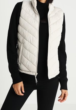 Lehká krémová vesta s prošíváním ve tvaru diamantů, vysokým límcem, předním zipem a bočními kapsami, doplněná černým svetrem.