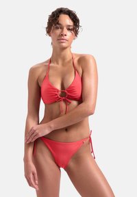 Czerwony bikini z trójkątnym stanikiem z centralnym detalem w formie pierścienia i regulowanymi troczkami, uzupełnione dopasowanymi, niskimi majtkami z bocznymi troczkami.