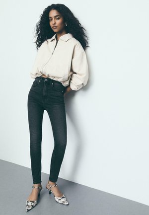 Frau mit langen lockigen Haaren, die eine beigefarbene Jacke mit Reißverschluss, schwarze enge Jeans und schwarz-weiß gemusterte Absätze trägt, steht vor einer weißen Wand.